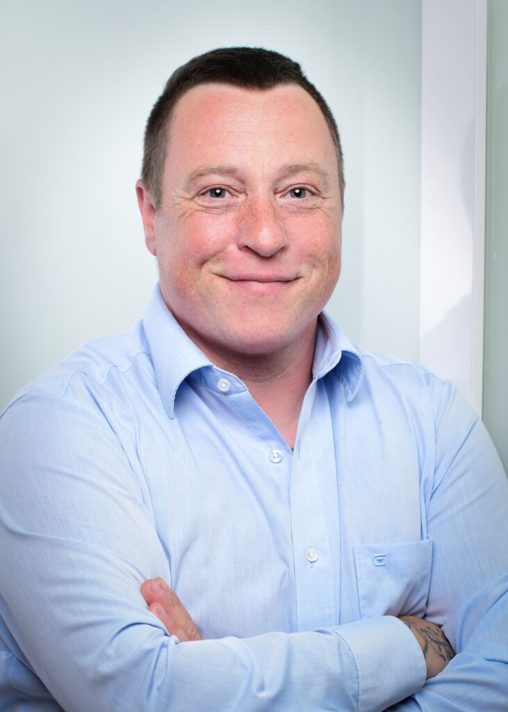 Bennet Kohl lächelt in die Kamera, er trägt ein hellblaues Hemd und hat die Arme locker verschränkt.