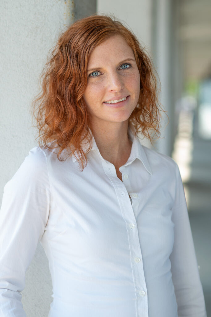 Jessica Schuh steht seitlich und lächelt in die Kamera. Sie trägt ihre roten Locken offen und hat eine weiße Bluse an.