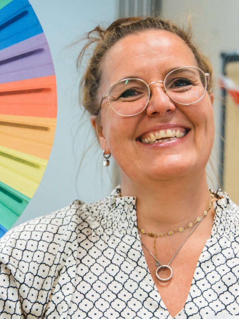  Kari Bischof-Schiefelbein strahlt in die Kamera. Sie trägt ihre Haare zusammen und hält einen Fächer in Regenbogenfarben am links seitlich ins Bild.