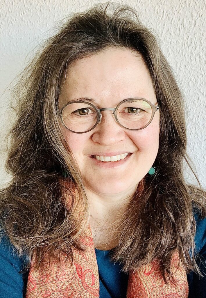 Solveig Chilla lächelt in die Kamera. Sie trägt eine Brille. Ihre dunkleblonden Haare offen und einen rotorangenen Schal.