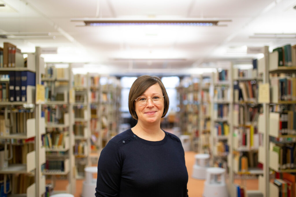 Nina Hogrebe ist in einer Bibliothek vor den Buchreihen zu sehen. Sie lächelt in die Kamera. Sie trägt eine Brille, ein dunkles Oberteil und ihre dunkelblonden Haare offen.