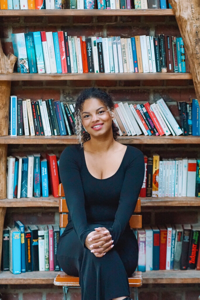 Chantal Chisley sitzt vor einem gut gefüllten Bücherregal. Sie hat ihre Hände auf ihre locker überschlagenen Beine gelegt und strahlt in die Kamera. Ihre Haare trägt sie so, dass die blonden Locken vorne aus dem Zopf fallen.