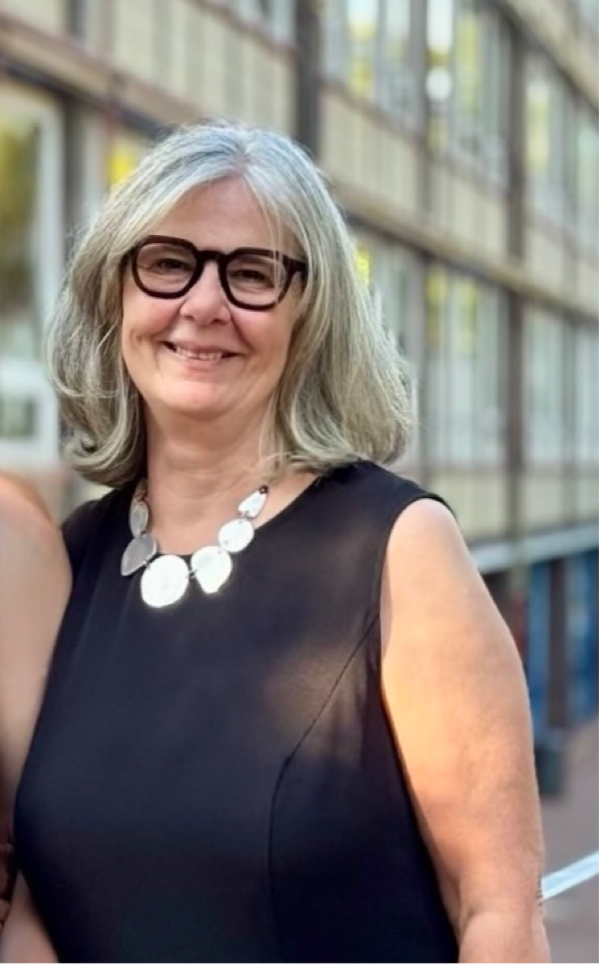 Frau Schaugg lächelt in die Kamera. Sie trägt eine Brille. Ihre hellen Haare fallen leicht auf das schwarze Kleid dass sie trägt.